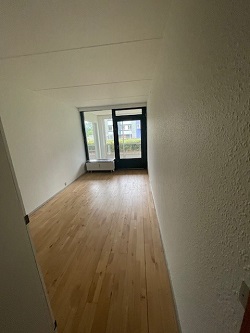 An empty room with light wooden flooring, white walls, and a large window and glass door providing natural light.