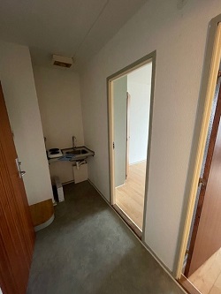 A narrow entrance/living area with a small sink and washing machine connection on the left, and an open doorway leading to another room on the right.