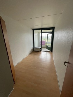An empty room with light wooden flooring, white walls, and a large glass door leading to a balcony.