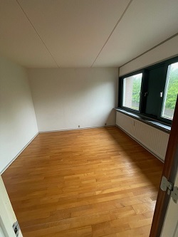 An empty room with wooden floorboards, white walls, and a large window with a radiator underneath, likely a bedroom.