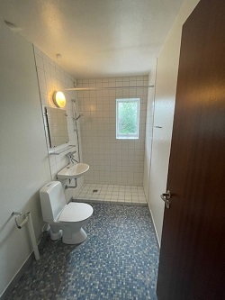 A bathroom with blue mosaic floor tiles, white tiled walls, a toilet, a small sink, and a shower area with a window.