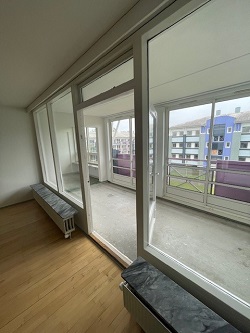 View from a room through large glass windows and a door onto an enclosed balcony, with a view of other apartment buildings.