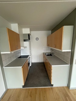 A galley kitchen with light wood cabinets, grey countertops, a stove, sink, and range hood.