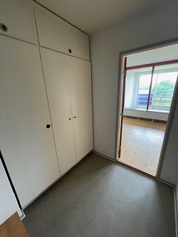 View of an entrance area with built-in white wardrobes and a doorway leading to a bright room with a large window.