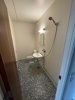 A compact bathroom with a toilet, sink, mirror, and shower head, featuring a speckled grey floor.
