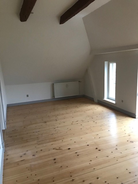A bright, unfurnished attic room with light wooden flooring, white walls, a sloped ceiling with exposed beams, a radiator, and a tall window.