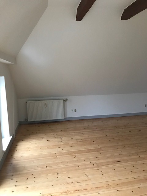A wide view of an unfurnished attic room with light wooden flooring, white walls, a sloped ceiling with exposed beams, and a radiator.