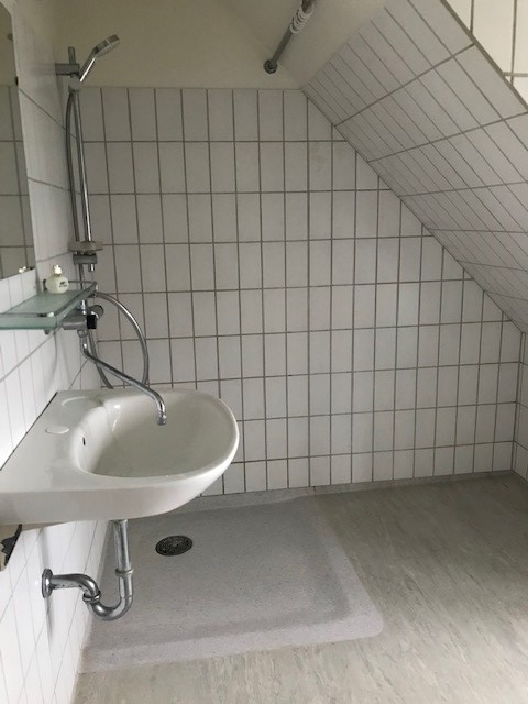 A compact bathroom with white tiled walls, a sloped ceiling, a wall-mounted sink, and a shower area with a shower head and a shower tray.