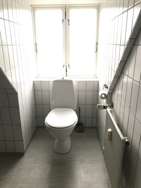 Bílá toaleta v úzké, obložené koupelně se šikmým stropem, oknem a radiátorem.