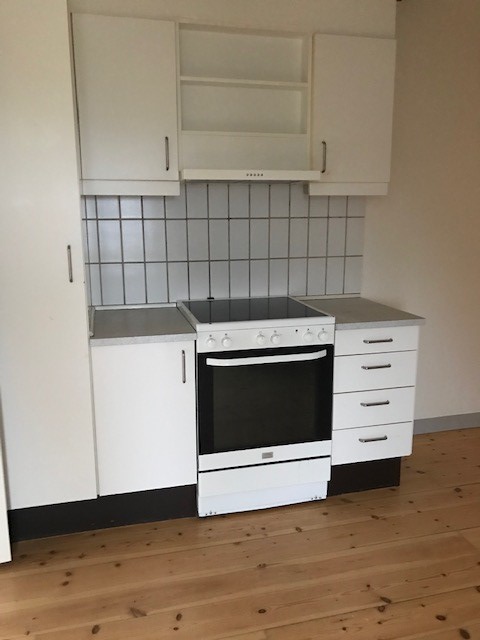 Kompaktní kuchyňský prostor s bílými skříňkami, integrovanou troubou a varnou deskou, odsavačem par a obkladem, na pozadí dřevěné podlahy.