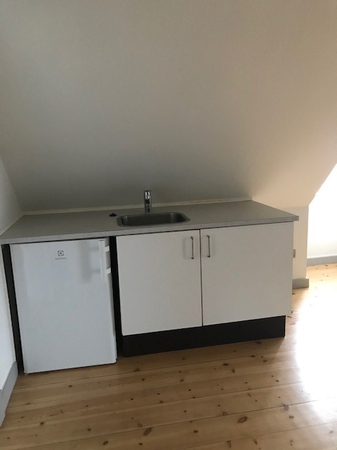 Malý kuchyňský kout s umyvadlem, lednicí pod linkou a bílými skříňkami, umístěný pod šikmým stropem s dřevěnou podlahou.