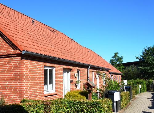 Side view of a red brick building with a tiled roof, showing windows, doors, and a birdhouse in the foreground, under a clear blue sky.