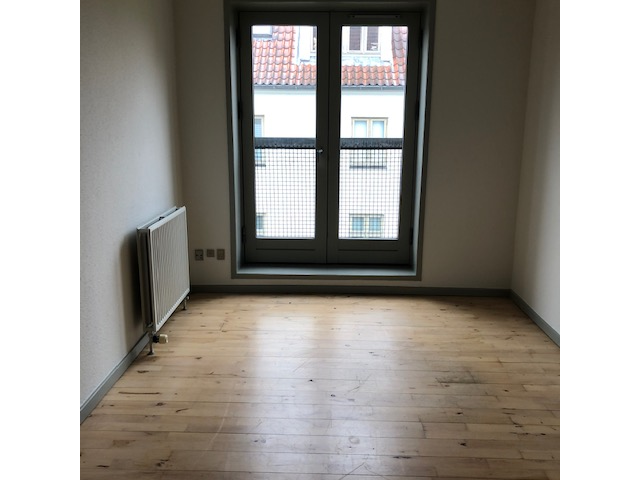 Seomra folamh le hurlár adhmaid éadrom, fuinneog mhór le greille, agus radaitheora ar an mballa ar chlé.