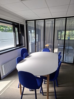 A meeting room with a white oval table and several blue chairs, next to large windows with dark curtains.