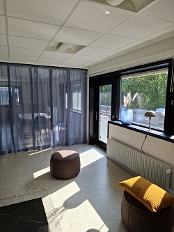 En lounge eller et fælles område med to brune osmanner, en gul pude og blå gennemsigtige gardiner, der dækker store vinduer.