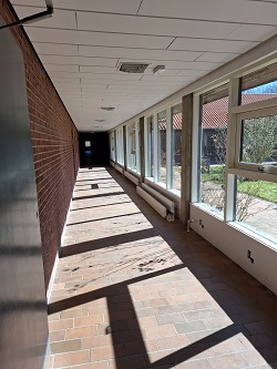 Long interior hallway with a brick wall on the left and large windows overlooking a courtyard on the right, with sunlight casting shadows on the floor.