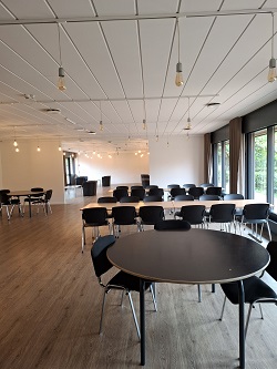 Large, modern multi-purpose room set up with various tables and chairs for meetings or dining, featuring light wood flooring and large windows.