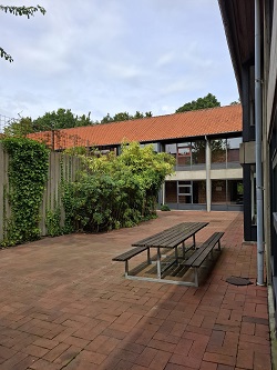 Exterior courtyard with a wooden picnic table and benches, surrounded by brick paving, green foliage, and a building with a red tile roof.