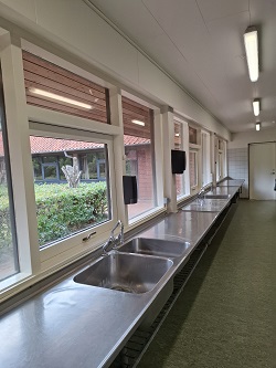 Lang kommerciel køkkenbord med flere vaske i rustfrit stål og store vinduer med udsigt over en gårdhave.