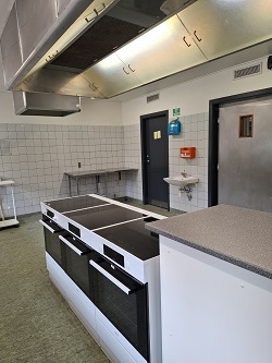 Kommercielt køkken med en stor central kogeø med flere ovne og komfurer, emhætte i rustfrit stål og flisebelagte vægge.