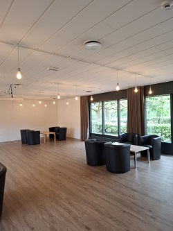 Udsnit af et stort, moderne multifunktionelt rum med lounge-siddeområder, med sorte lænestole, sofaborde og store vinduer.