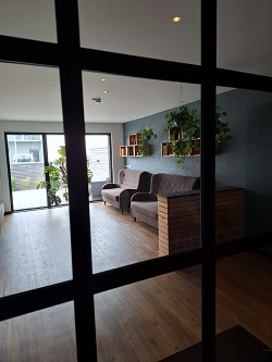 A modern lounge area with two grey sofas, wooden shelves with plants on a dark blue wall, and large glass doors leading outside.