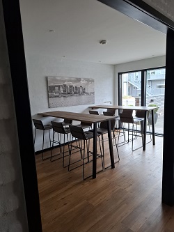 A communal dining or workspace area with long wooden high-top tables and bar stools, and a large window overlooking an outdoor area.