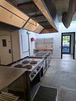 A professional commercial kitchen with large stainless steel cooking ranges, overhead ventilation hoods, and white tiled walls.
