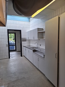 Another view of a commercial kitchen area with white tiled walls, stainless steel countertops, a sink, and white cabinets.