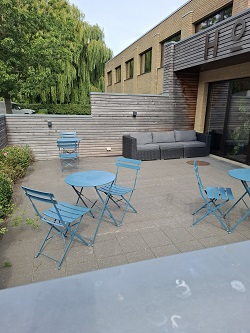 Limistéar patio lasmuigh le táblaí agus cathaoireacha miotail gorma, agus tolg liath lasmuigh suite i gcoinne fál adhmaid.