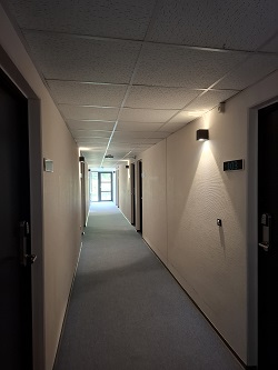 Dlouhá úzká chodba se světlými stěnami, šedým kobercem a několika dveřmi vedoucími do pokojů, osvětlená nástěnnými světly.