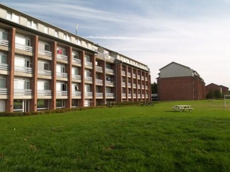 Exterior view of a multi-story residential building with balconies and a large grassy area in front, with another brick building in the background.