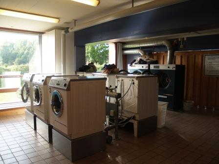 An interior view of a communal laundry room with several washing machines and dryers, large windows, and tiled floors.