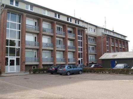 Přední exteriér vícepodlažní obytné budovy s velkými okny, balkony a parkovištěm s auty v popředí.