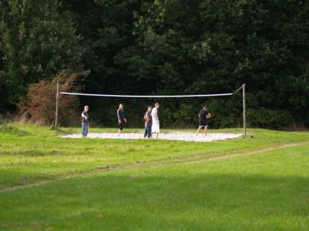 A group of people playing volleyball on a sand court with a net, surrounded by green grass and trees.
