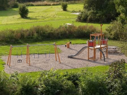 An aerial view of a children's playground with swings and a slide structure on a sandy base, surrounded by green fields and trees.