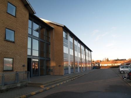 Vnější pohled na cihlový bytový dům s velkými okny a parkovištěm pod jasně modrou oblohou.