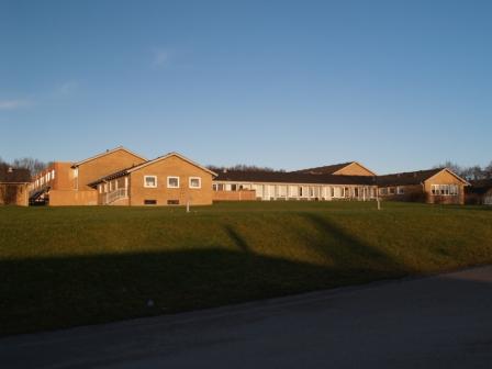 Välivaade pikale madalale pruunika katusega telliskivihoonele, mis asub selge sinise taeva all rohusel nõlval.