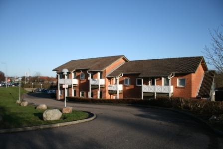 Wide shot of a brick residential building with multiple balconies, a curved driveway, and landscaping under a clear blue sky.