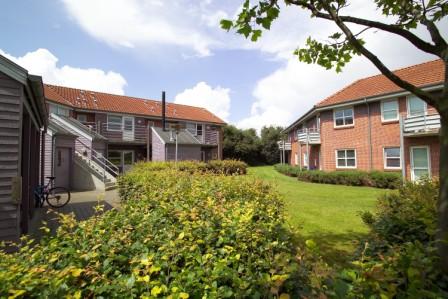Courtyard view between two residential buildings with red tile roofs, featuring a green lawn and bushes under a partly cloudy sky.